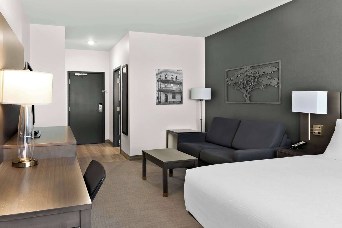 A hotel room with a bed, a desk, a chair, a sofa, a small table, and a lamp.