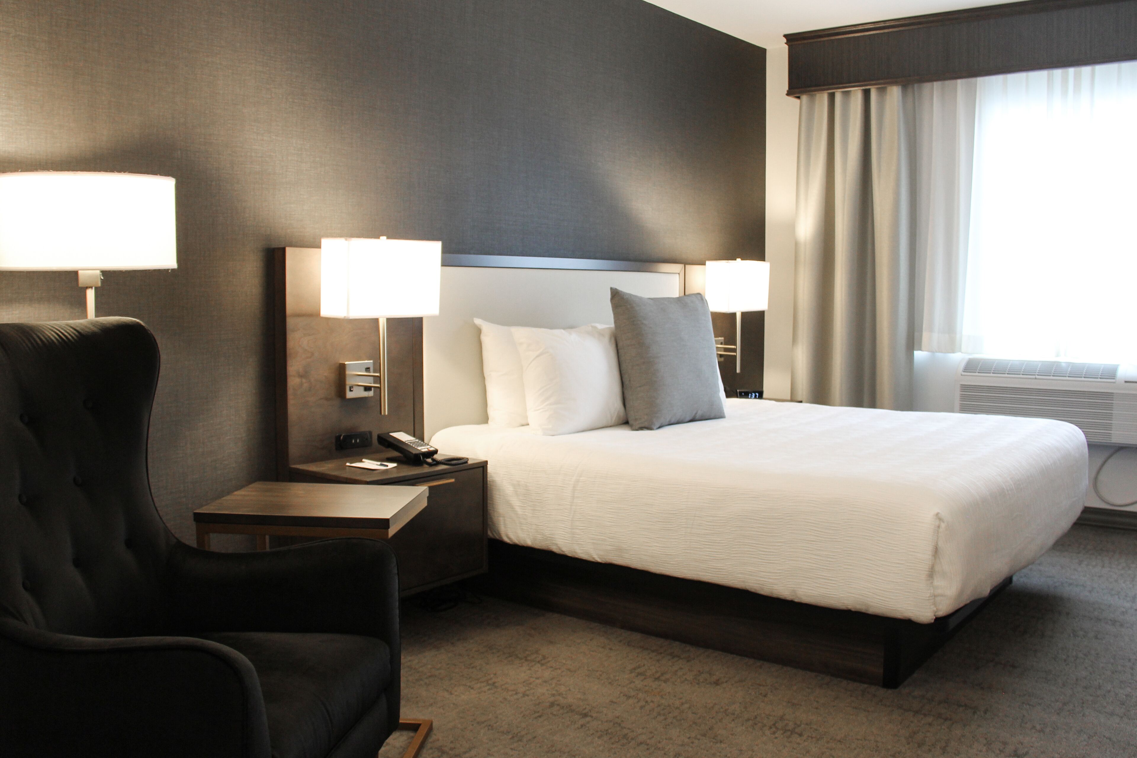 A hotel room with a large bed, two bedside tables with lamps, a chair, and a window with curtains.