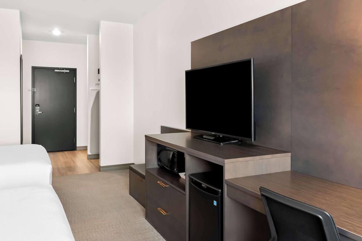 A modern hotel room with a bed, a desk with a flat-screen TV, a microwave, and a refrigerator.