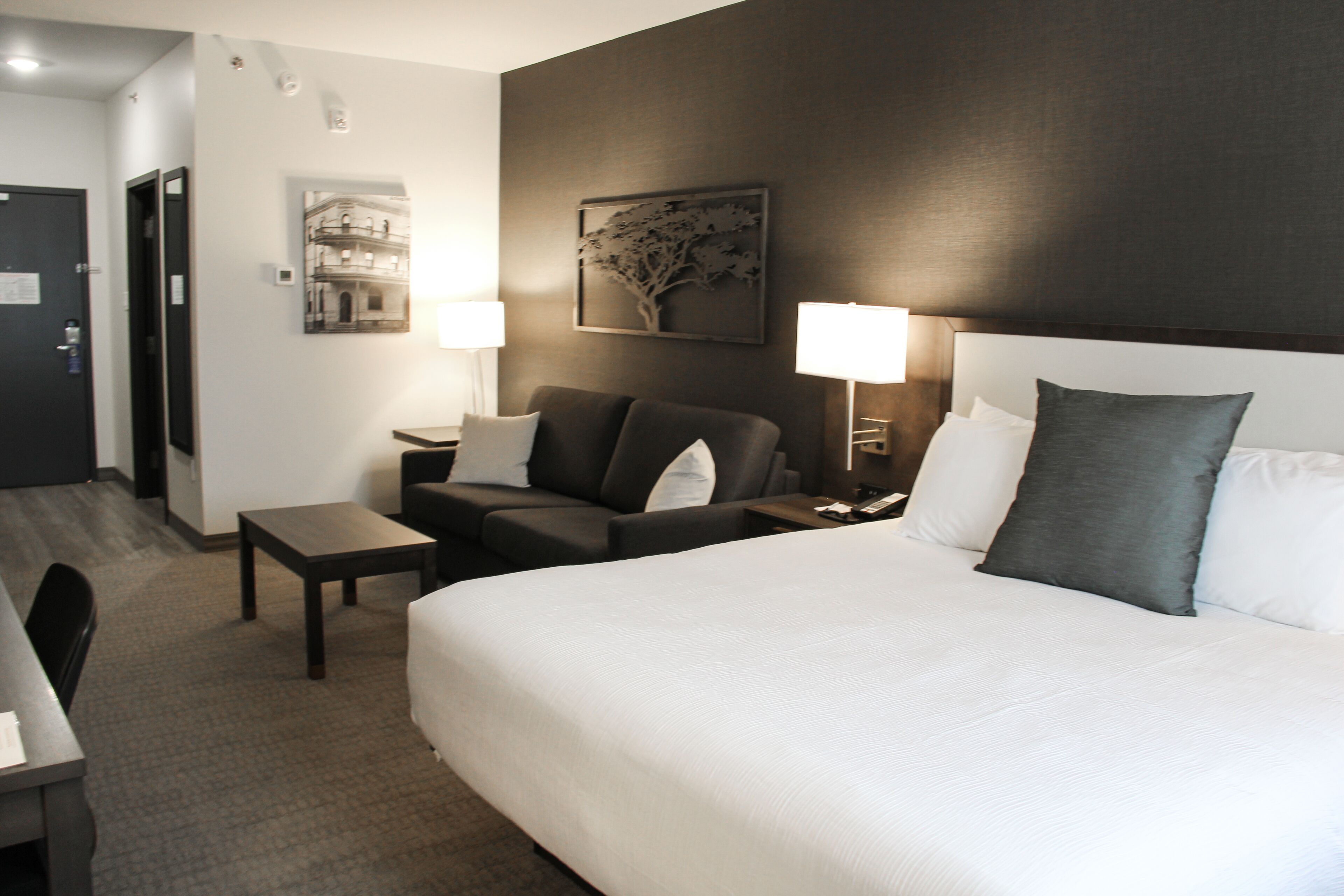 A hotel room with a large bed, a desk, a chair, two lamps, and a wall-mounted painting.