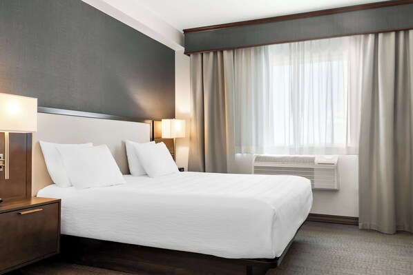 A hotel room with a large bed, two bedside lamps, a nightstand, a window with curtains, and an air conditioning unit. - Best Western Plus Morden (Morden)