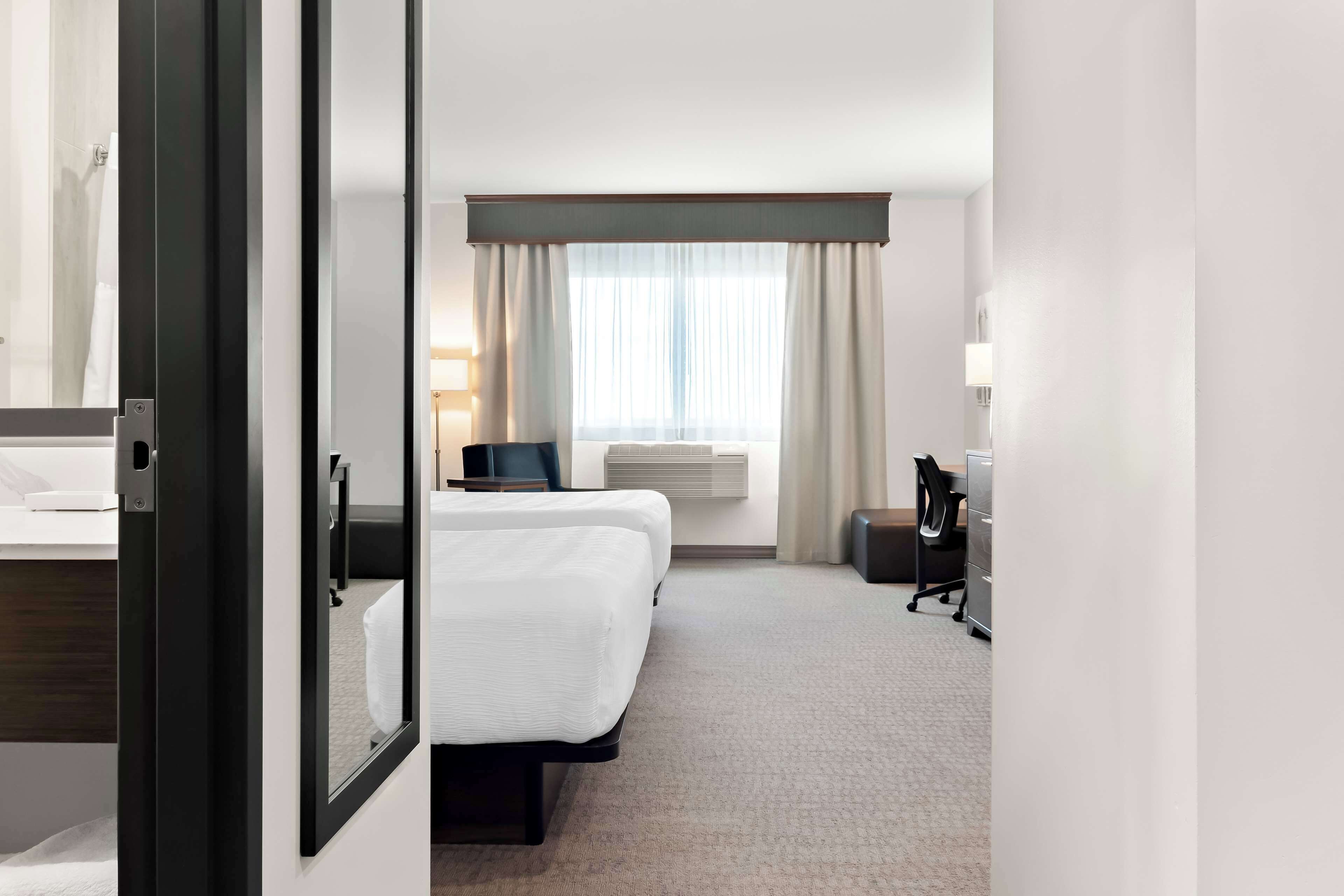 Una habitación de hotel con cama, escritorio, silla, lámpara y ventana con cortinas.