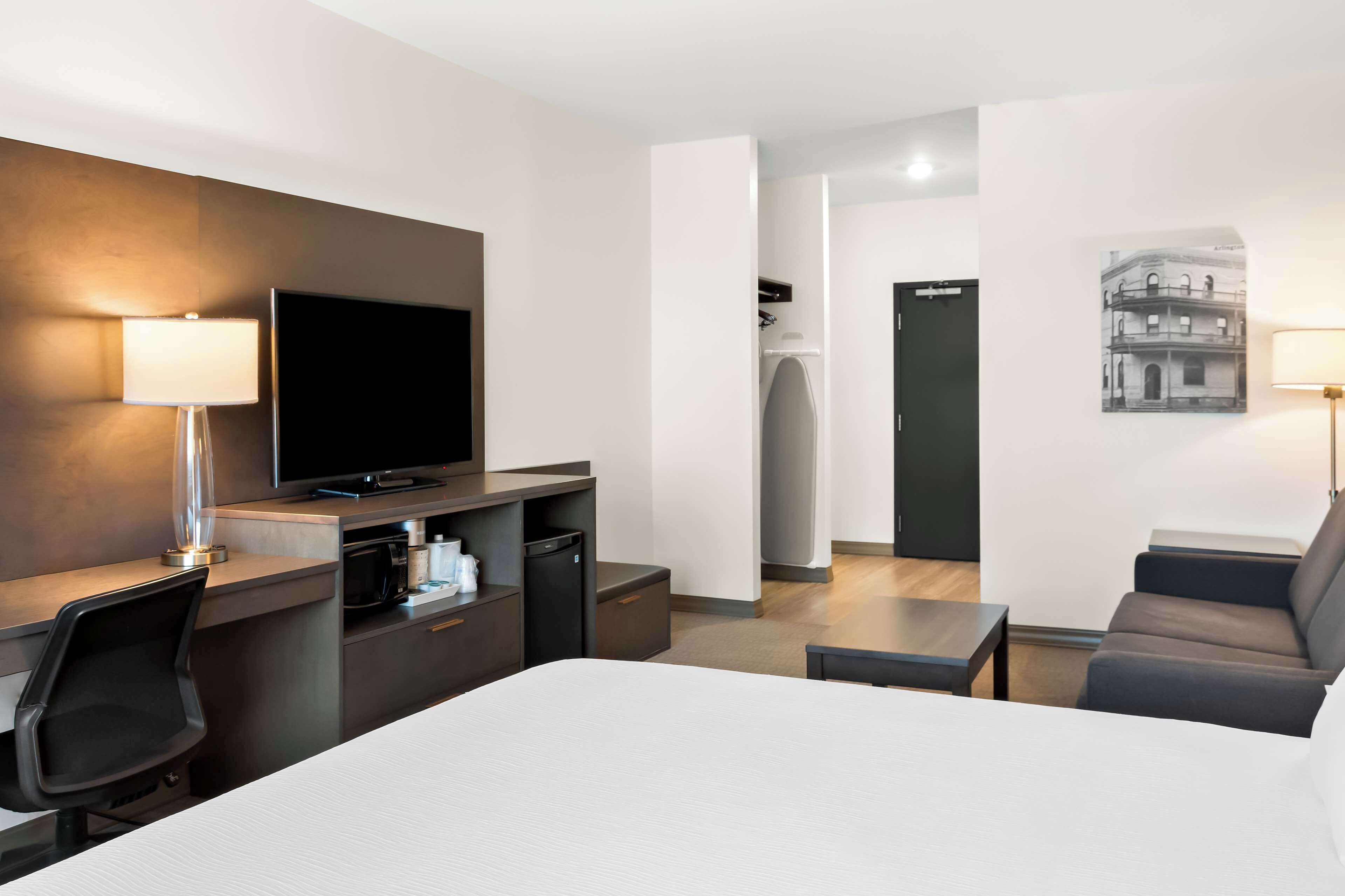 A hotel room with a bed, a desk with a computer, a TV, a sofa, and a bathroom visible through an open door.