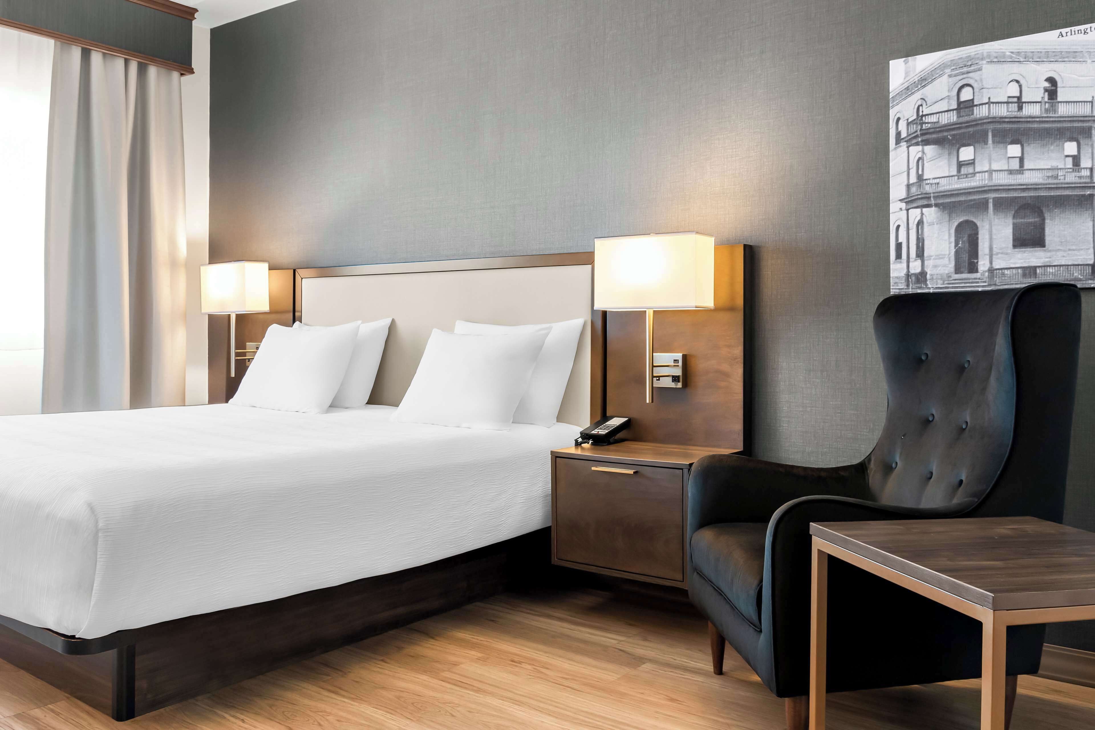 A hotel room with a large bed, two bedside lamps, a nightstand, a chair, and a small table.