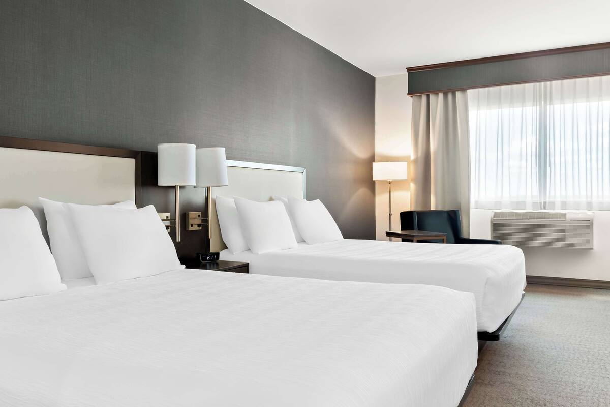 A hotel room with two beds, a nightstand with a lamp, a chair, and a large window with curtains.