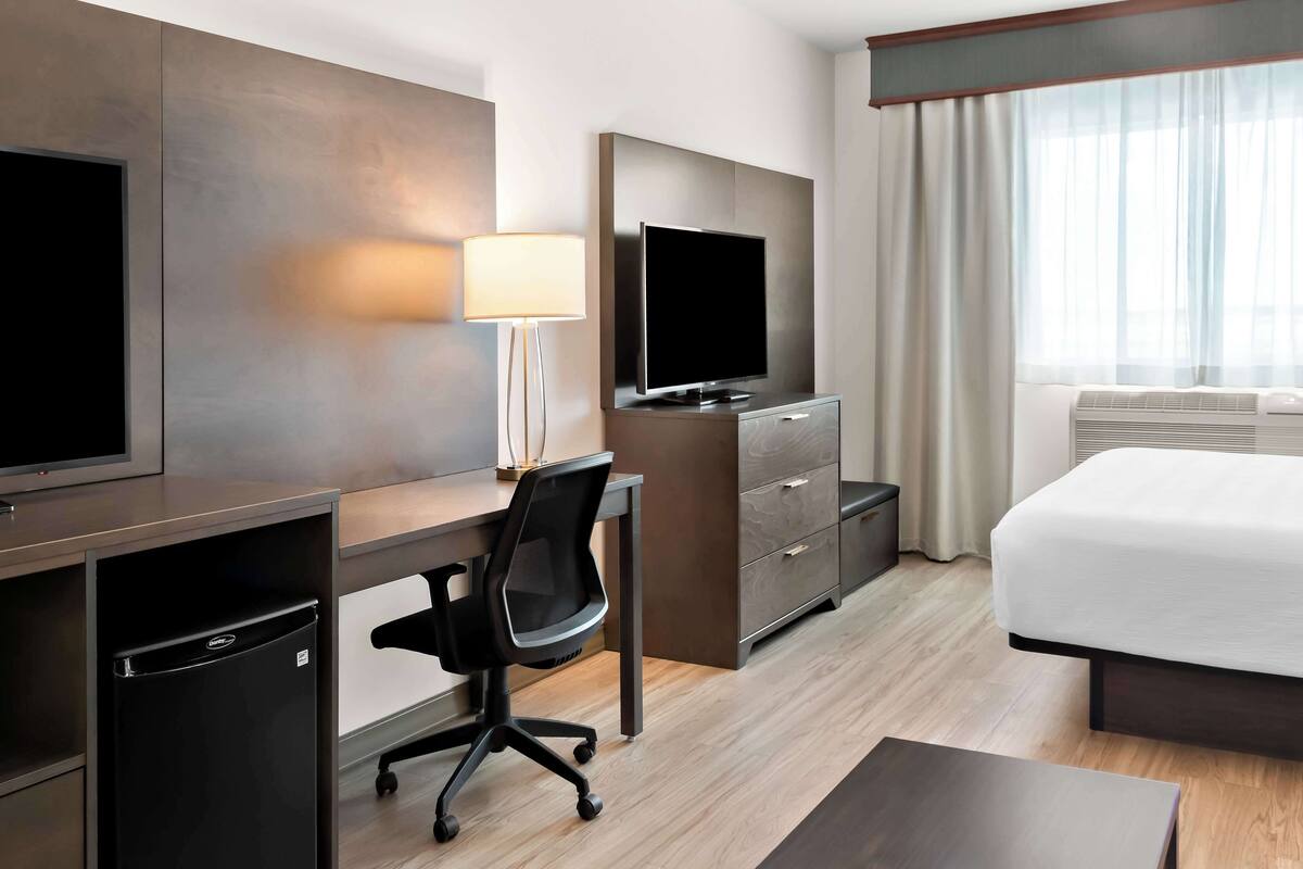 A hotel room with a bed, a desk with a computer, a TV, a lamp, and a minibar.
