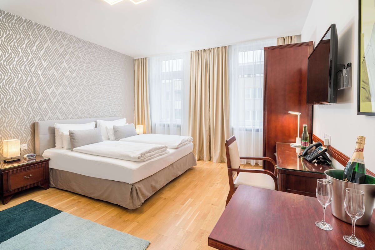 A hotel room with a bed, bedside table, lamp, and a desk with a chair and a bucket of champagne glasses.