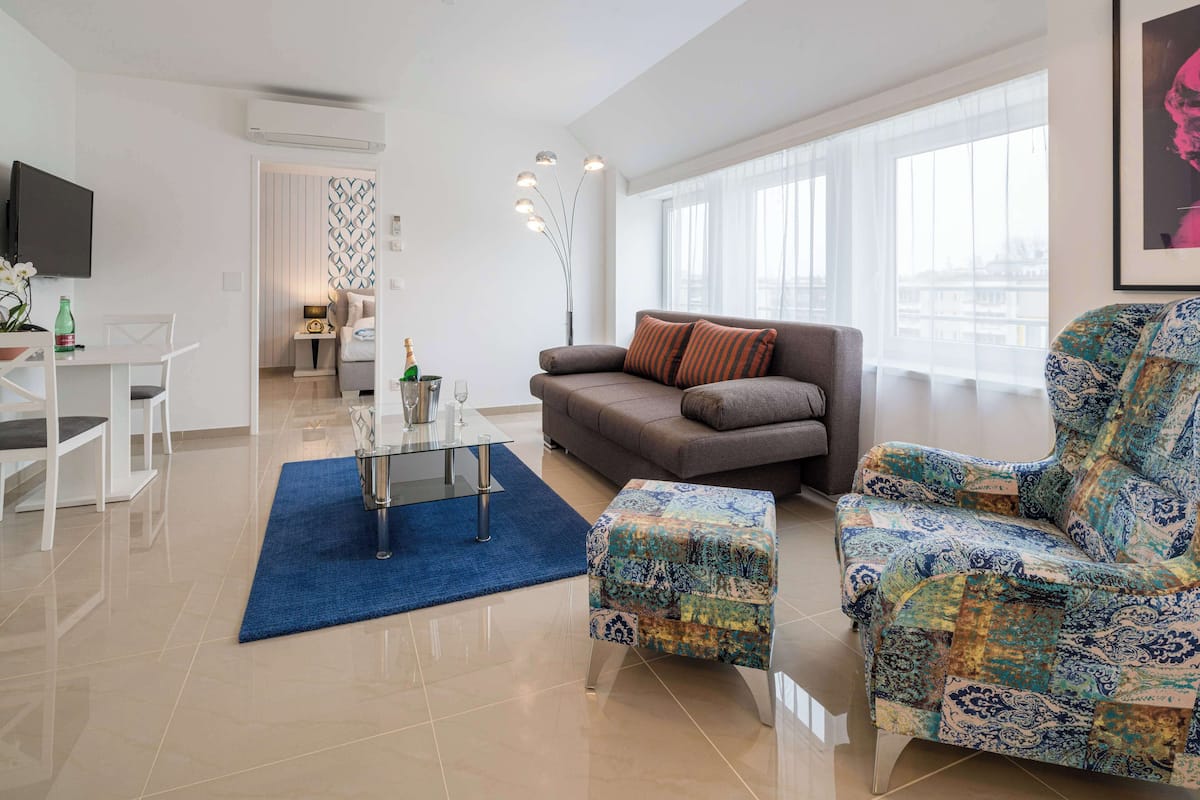 A modern living room with a sofa, ottoman, and coffee table. There is a TV on the wall, a dining area with chairs, and a patterned rug.