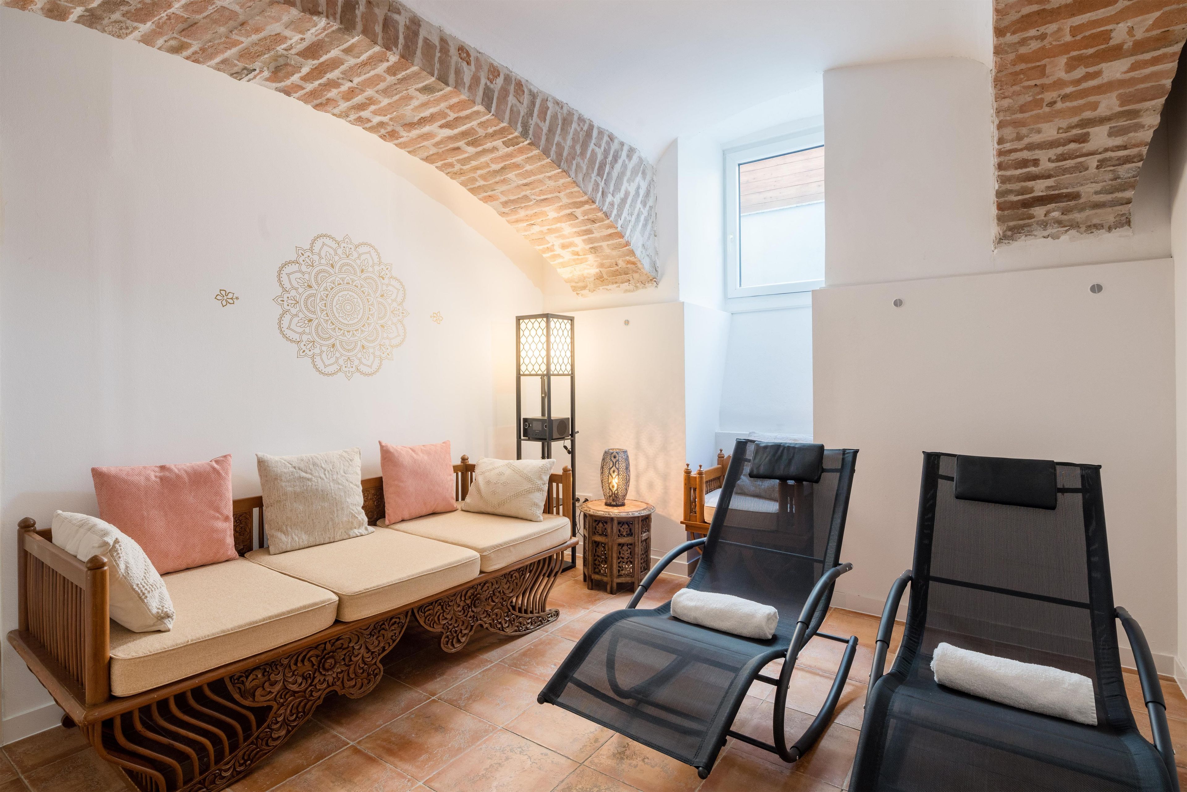 A room with a brick arch ceiling, a sofa with cushions, a chair with a towel, a small table with a lamp, and a decorative wall piece.
