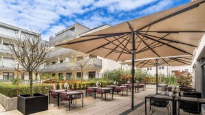 An outdoor patio area with multiple tables and chairs, large umbrellas, and a view of residential buildings in the background. - Aiden by Best Western Biberach (Biberach an der Riss)