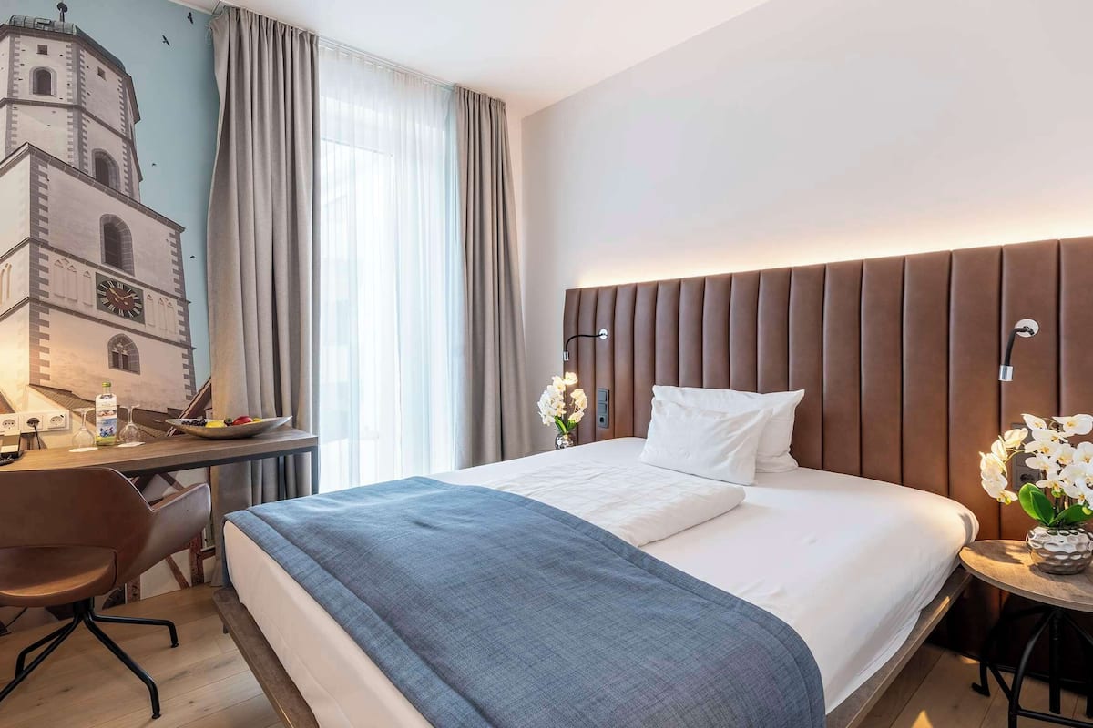 Habitación de hotel con cama, escritorio, silla, una pequeña mesa con flores y un mural en la pared.