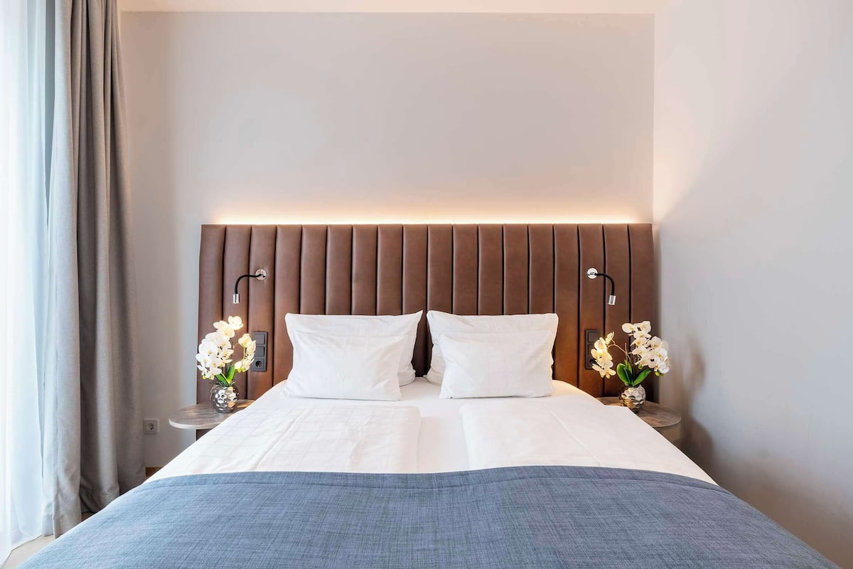 Una cama bien tendida con ropa de cama blanca, un cabecero de madera y dos mesitas de noche con lámparas y jarrones de flores.