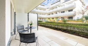 A modern outdoor patio with two black metal chairs and a small round table, a white railing, and a view of a multi-story building with balconies. - Aiden by Best Western Biberach (Biberach an der Riss)