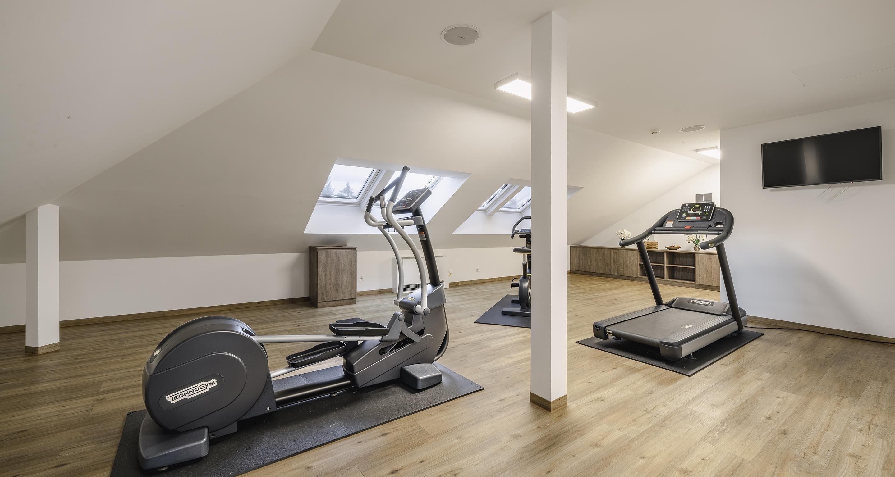 A modern gym with two elliptical machines, a treadmill, and a TV on the wall.