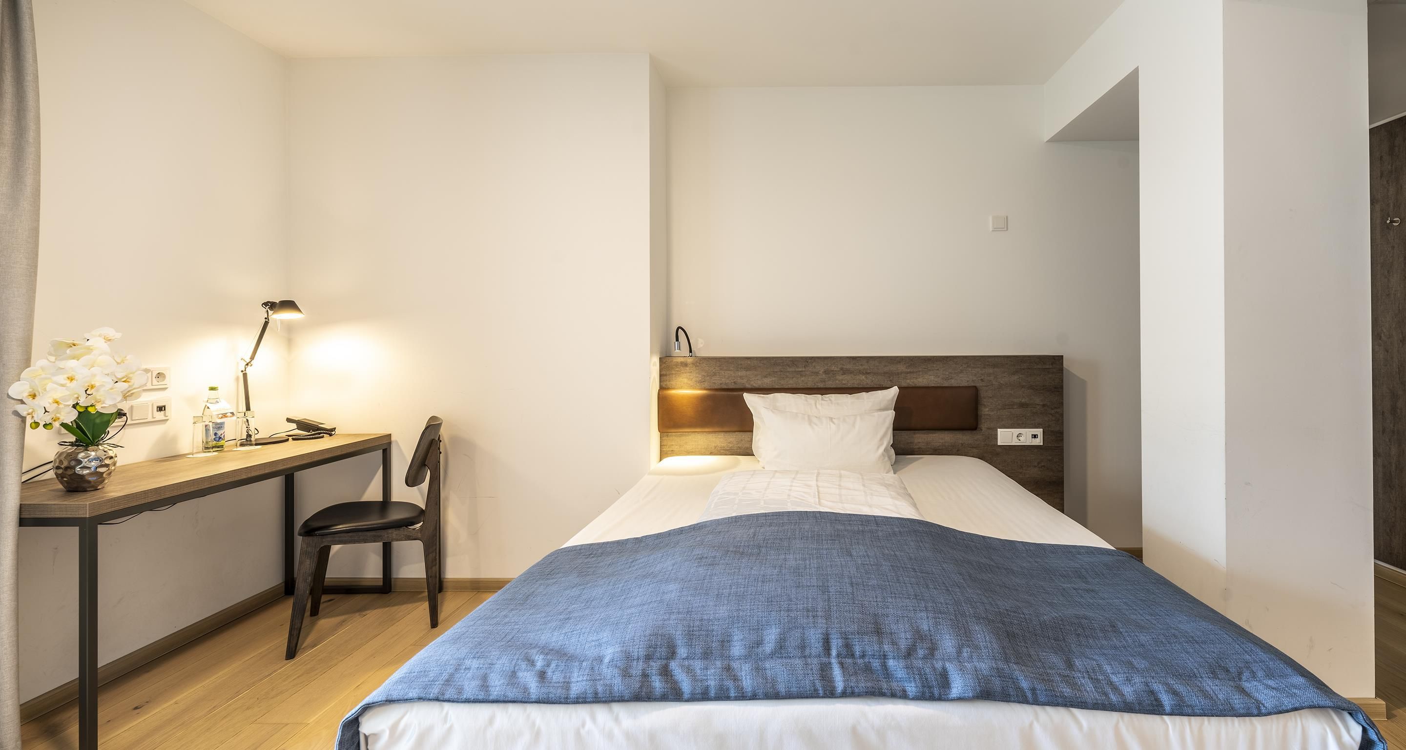Habitación de hotel con cama, mesita de noche, lámpara, silla y escritorio con lámpara y flores.