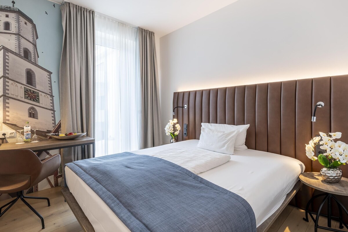 A hotel room with a bed, a desk, a chair, a small table with flowers, and a wall mural.