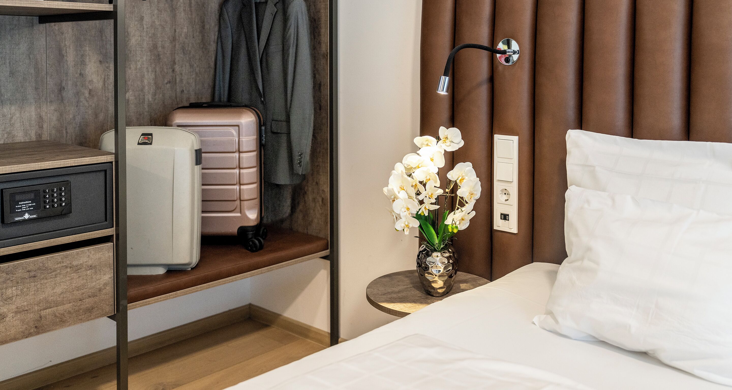 A hotel room with a bed, a bedside table with a vase of flowers, a suitcase, a coat rack, and a desk with a computer and a telephone.