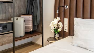A hotel room with a bed, a bedside table with a vase of flowers, a suitcase, a coat rack, and a desk with a computer and a telephone.