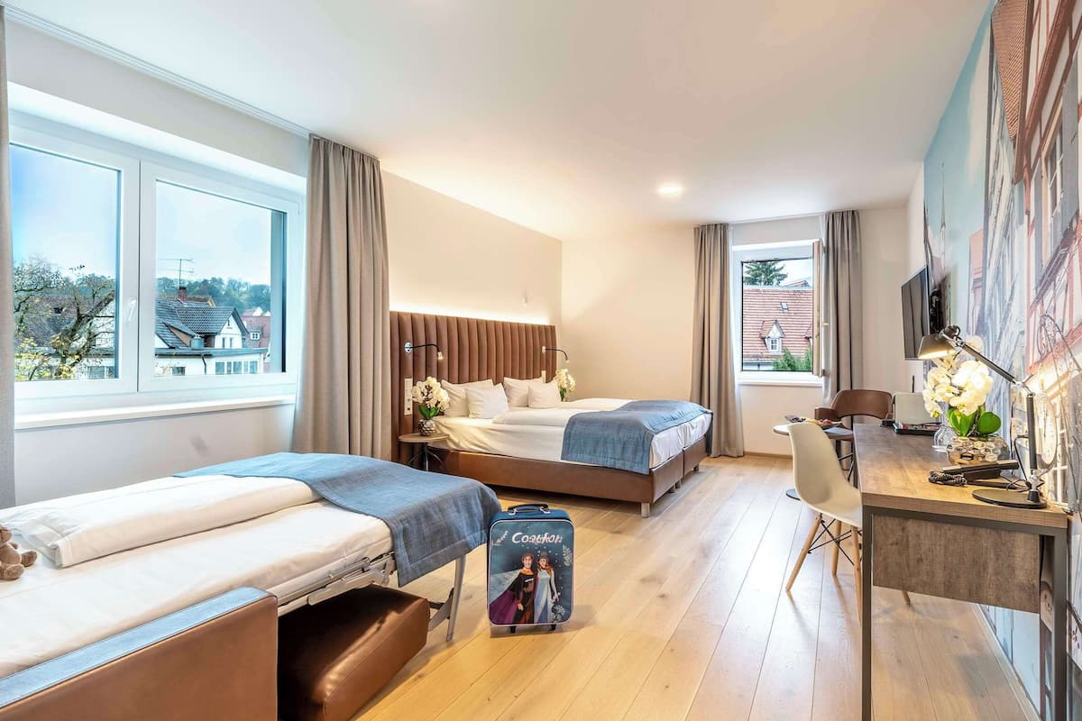Una habitación de hotel con dos camas, un escritorio con silla, una maleta y un televisor en la pared.