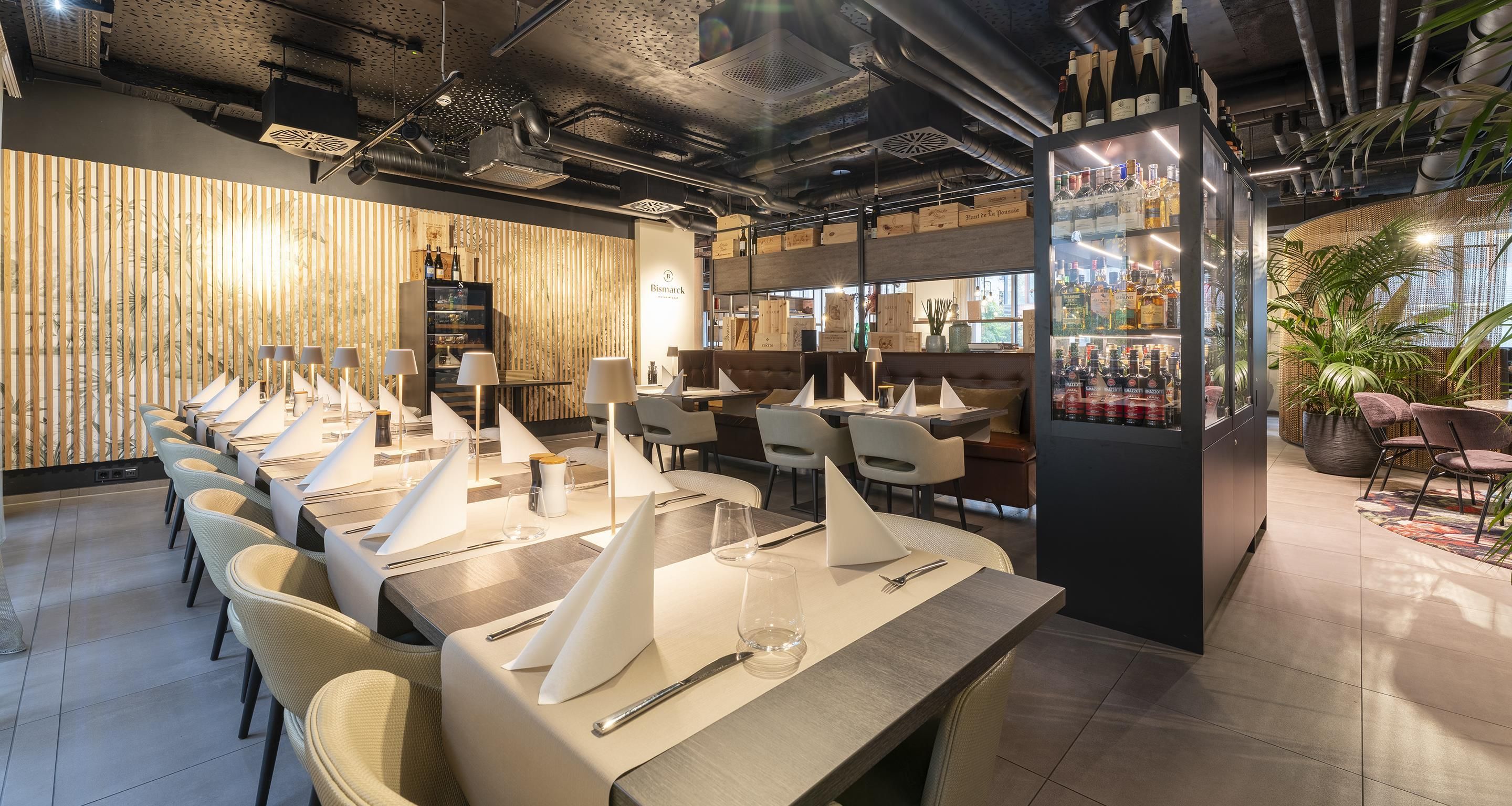 A modern restaurant interior with a long dining table, chairs, and a bar area with bottles.