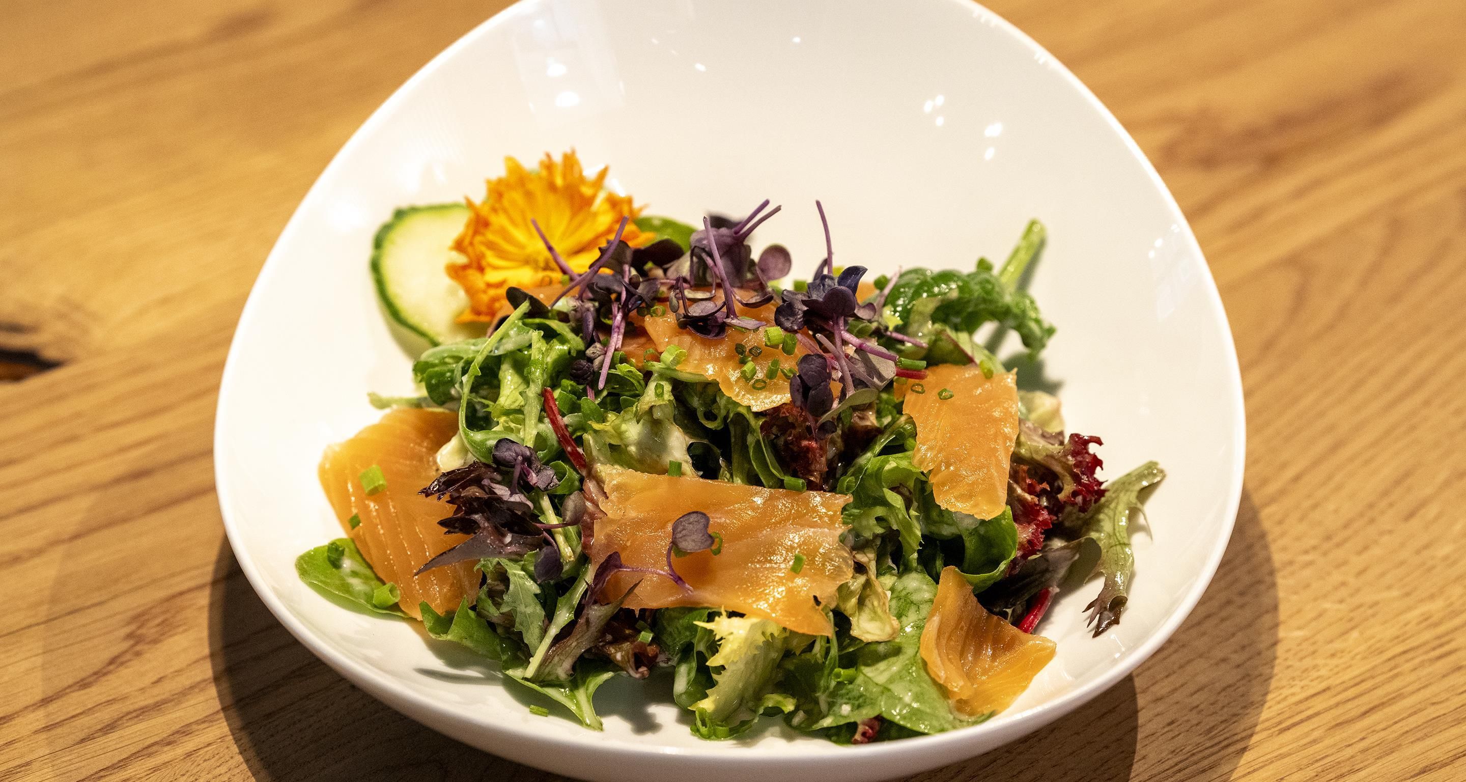 A white bowl containing a salad with various greens, a slice of salmon, and a decorative flower.