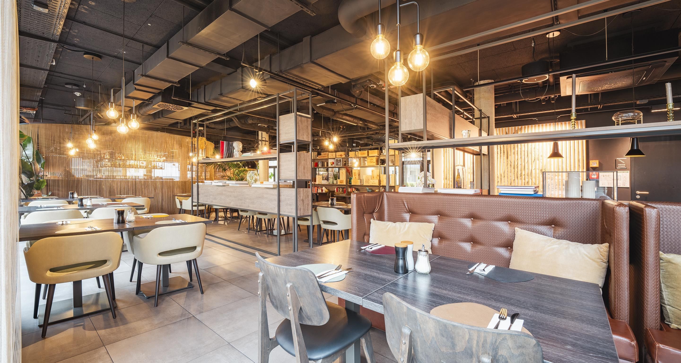 Interior modern d'un restaurant amb sostres alts, conductes a la vista i il·luminació penjant.
