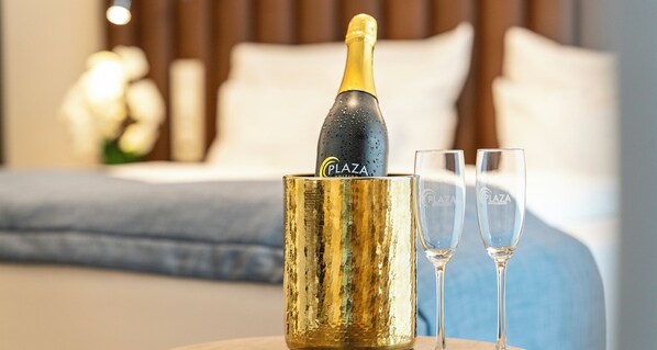 A champagne bottle in a gold container with the word "PLAZA" on it, two empty glasses, and a bed with a blue headboard in the background.