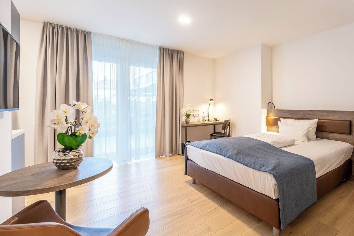 Una habitación de hotel moderna con una cama grande, un escritorio con lámpara, una silla y un florero con flores.