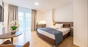 A modern hotel room with a large bed, a desk with a lamp, a chair, and a vase of flowers.