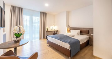 Quarto de hotel moderno com uma cama grande, uma escrivaninha com luminária, uma cadeira e um vaso de flores.