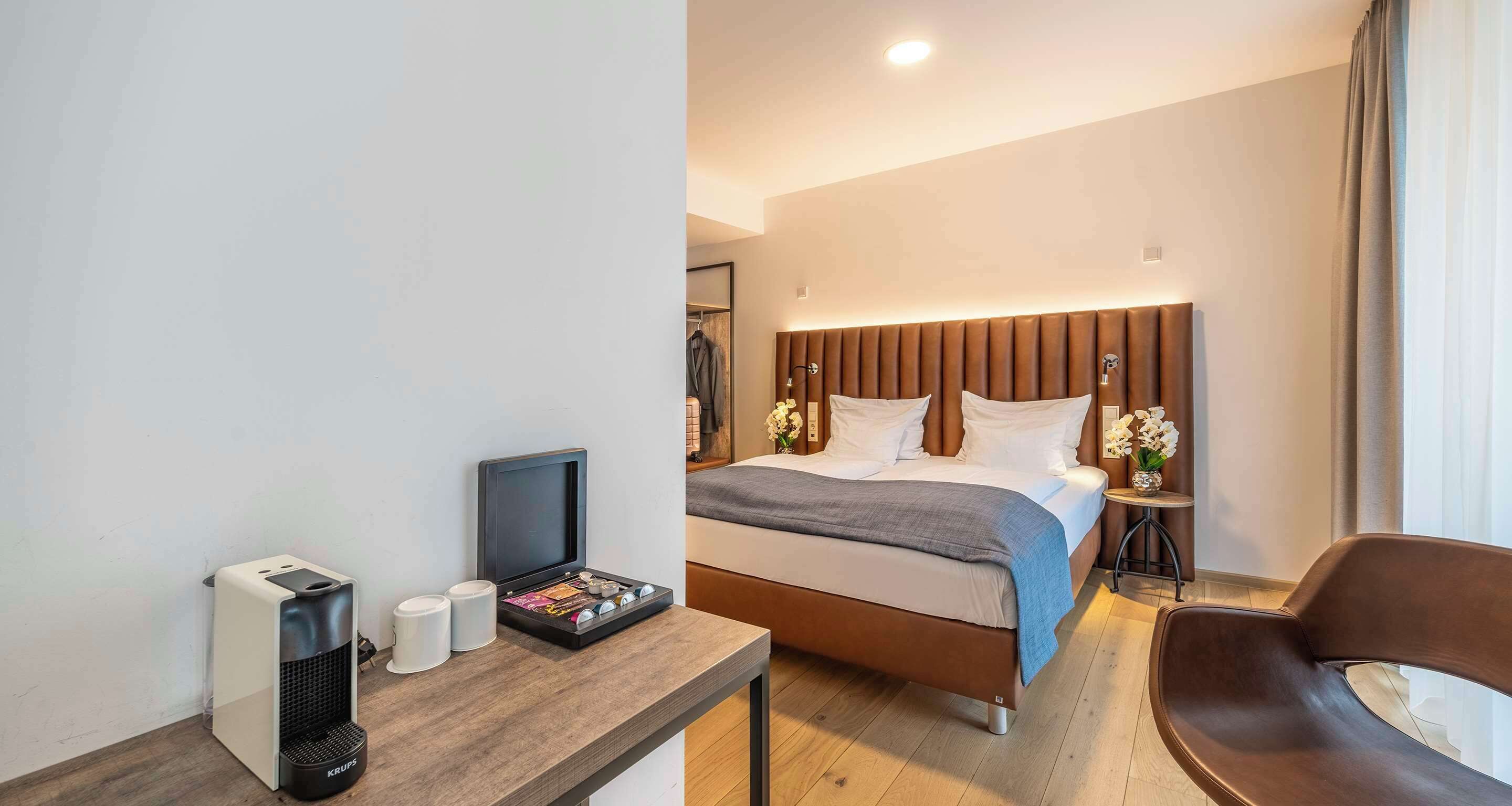A modern hotel room with a bed, bedside tables, a desk with a coffee machine, and a chair.