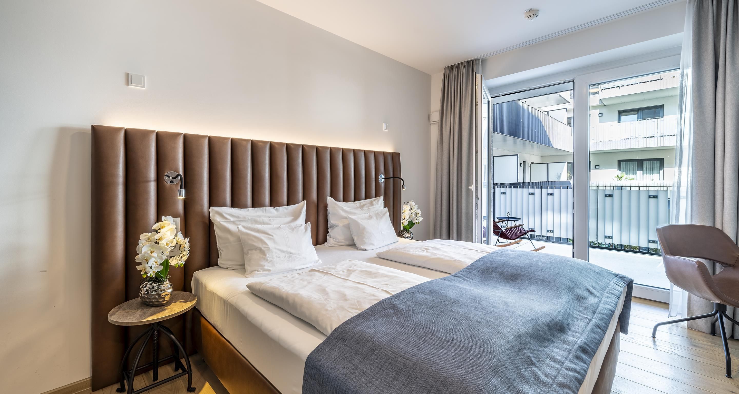Ein Hotelzimmer mit einem großen Bett, einem kleinen runden Tisch mit Blumen und Blick auf einen Balkon mit Gartenmöbeln.