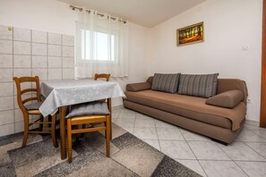 Uma sala de estar com um sofá marrom, uma mesa de jantar com duas cadeiras e uma janela com cortinas leves.