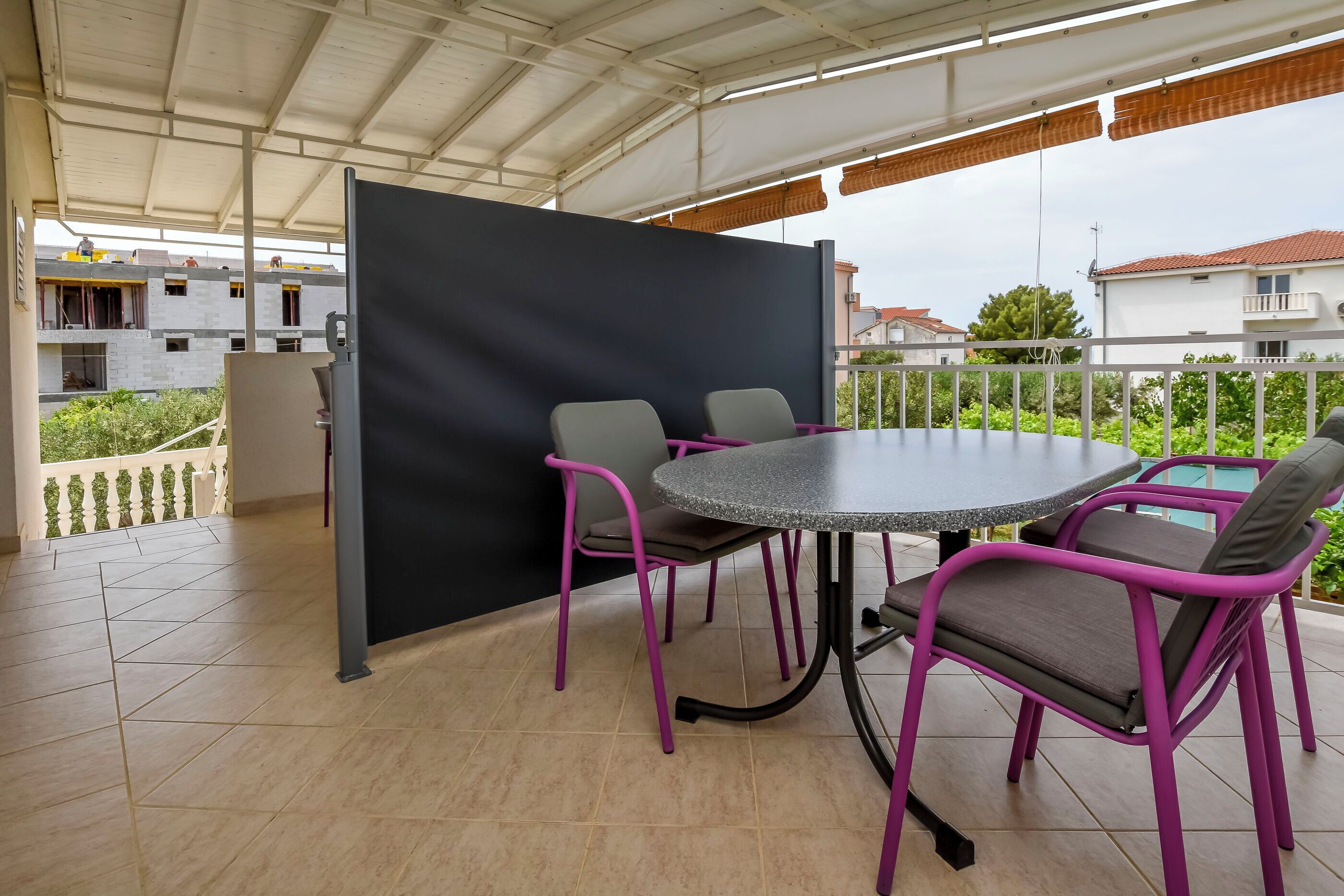 A covered outdoor area with a round table and four chairs, a black partition, and a view of buildings in the background.