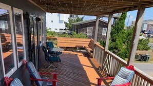 A wooden deck with red and blue Adirondack chairs, a table, and a covered area with a wooden railing. - Ocean Views at Cochran’s Corner! (Kill Devil Hills)
