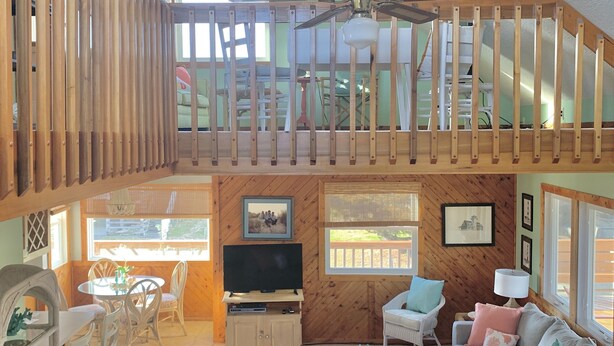 A spacious living area with a high ceiling, a wooden staircase, a television, and a sofa. - Ocean Views at Cochran’s Corner! (Kill Devil Hills)