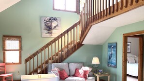 A cozy living room with a staircase, a couch, a table, and a painting of a boat. - Ocean Views at Cochran’s Corner! (Kill Devil Hills)