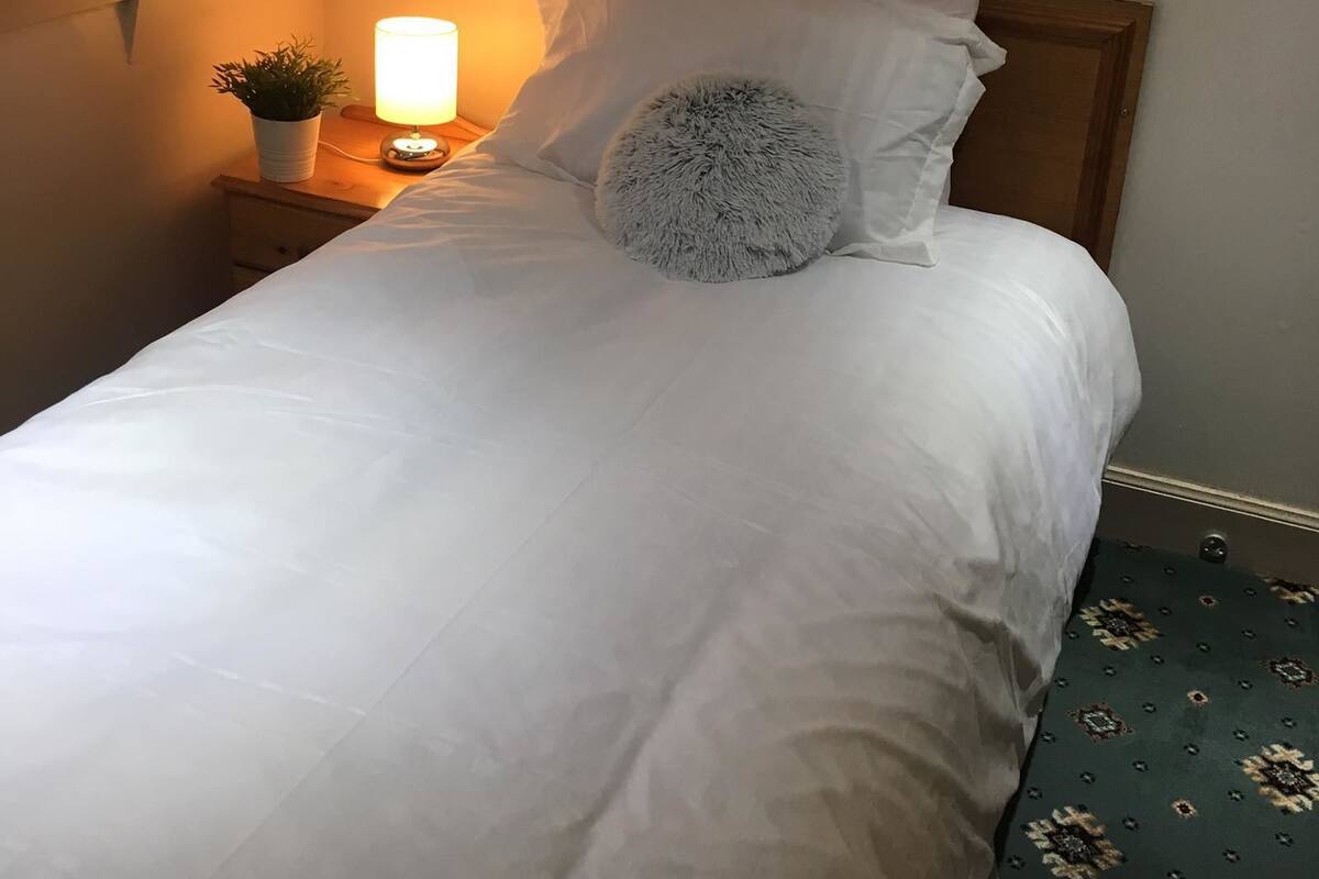 A neatly made bed with white linens, a fluffy pillow, and a bedside lamp on a wooden nightstand.