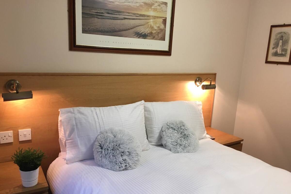 A neatly made bed with white linens, two fluffy pillows, a framed landscape painting above, and bedside lamps on wooden nightstands.
