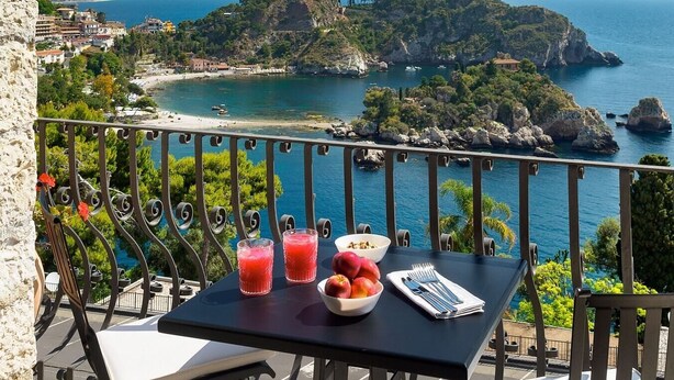 Un balcón con una mesa dispuesta para dos, con vista a un paisaje costero con afloramientos rocosos y una playa.