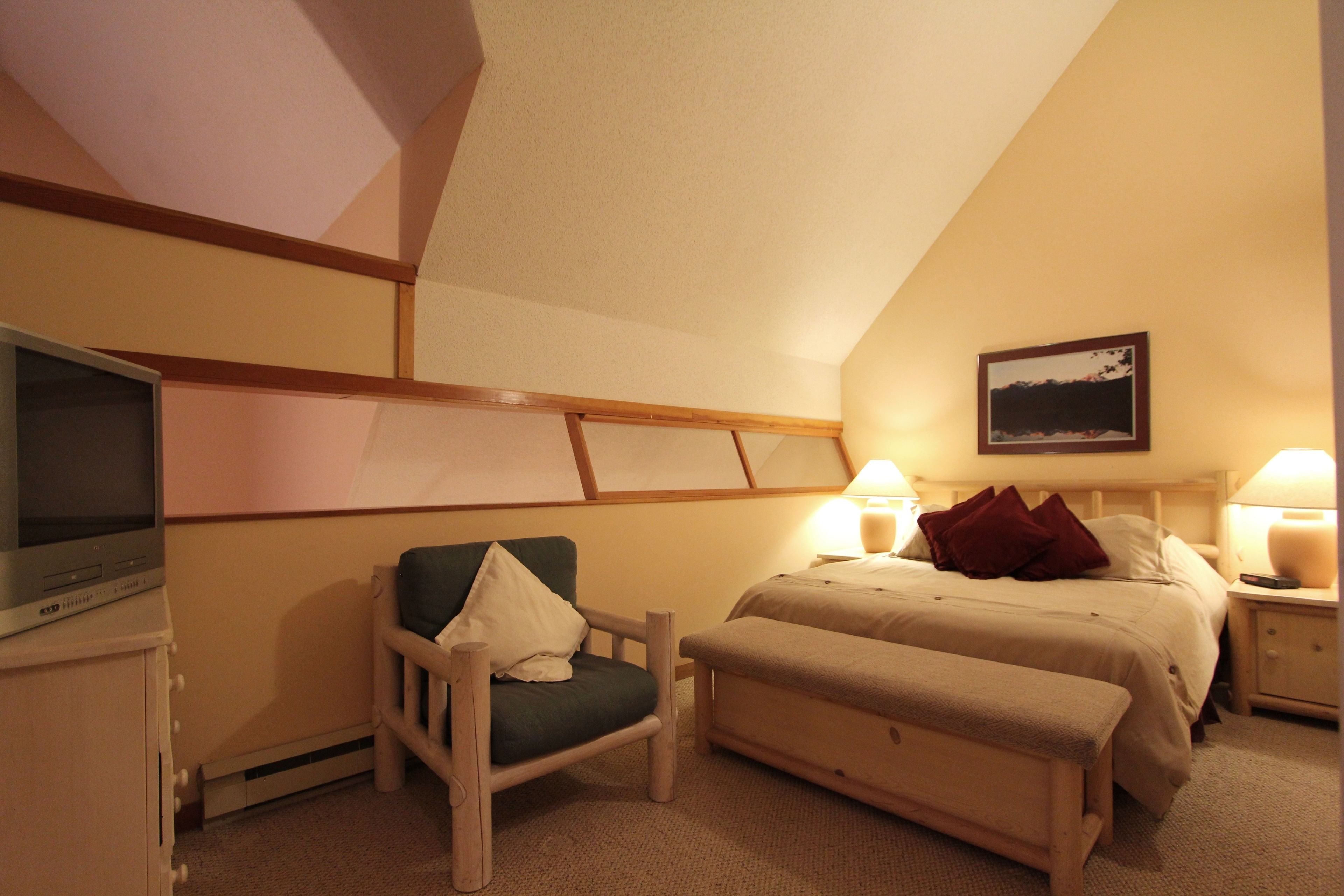 A hotel room with a bed, a chair, a bench, a television, and a nightstand with lamps.