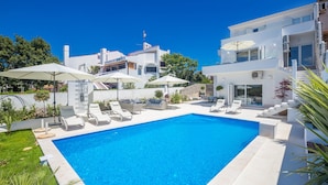 A modern pool area with lounge chairs and umbrellas, surrounded by white buildings and greenery. - Villa Andrea (Punat)