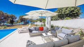 An outdoor seating area with wicker furniture, a pool, and umbrellas providing shade. - Villa Andrea (Punat)