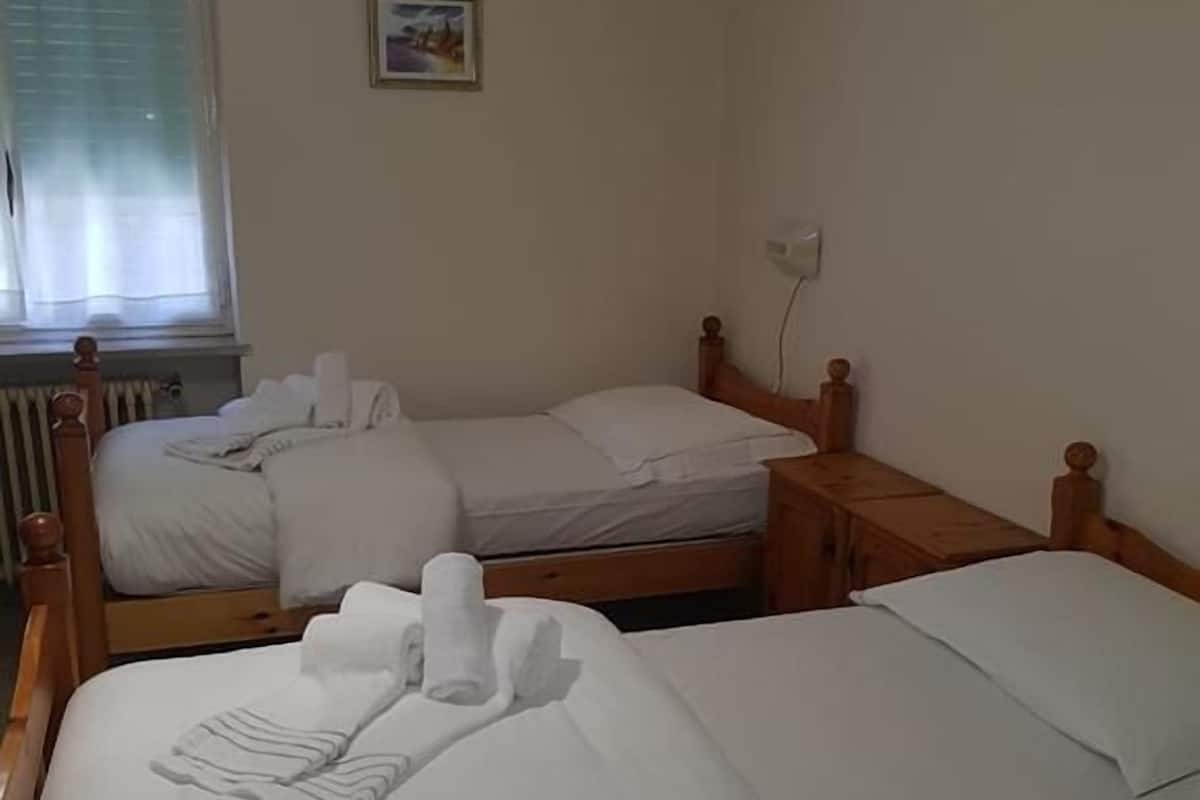 Quarto de hotel com duas camas de madeira, cada uma com roupa de cama e toalhas brancas. Há um pequeno quadro na parede e uma janela com persianas.