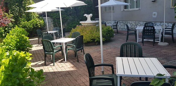 An outdoor patio area with tables and chairs, surrounded by greenery and a stone wall.
