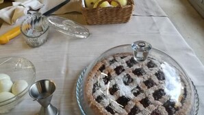A table with various food items, including a covered cake, a basket of apples, and a bowl of eggs.