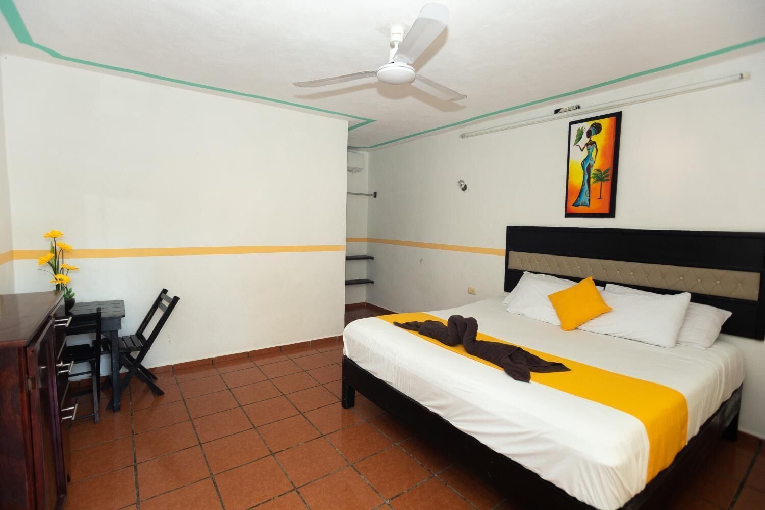 A hotel room with a bed, a desk with chairs, a ceiling fan, and a painting on the wall.
