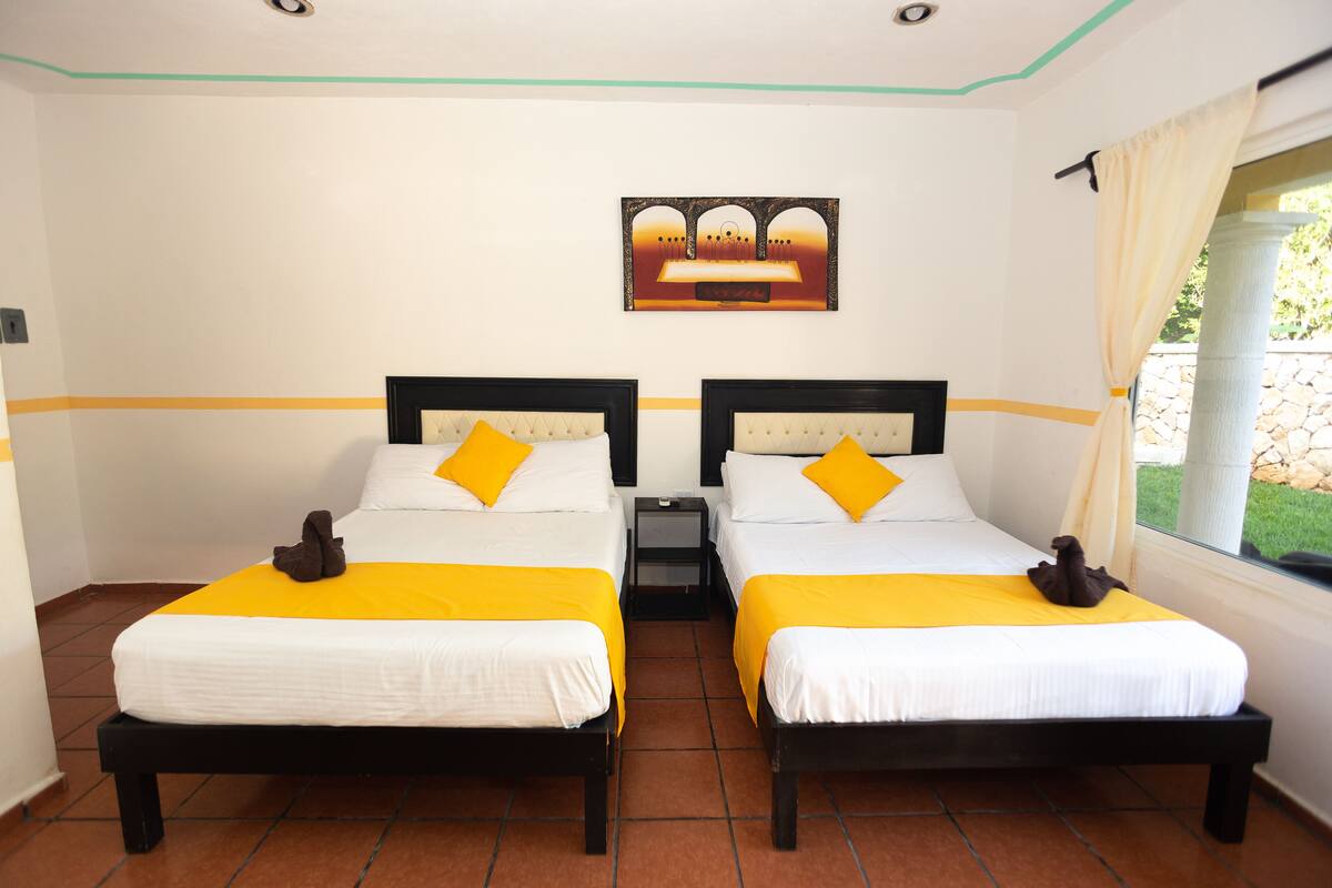 Habitación de hotel con dos camas, cada una con colchón blanco y colcha amarilla, y una mesita de noche entre ellas.