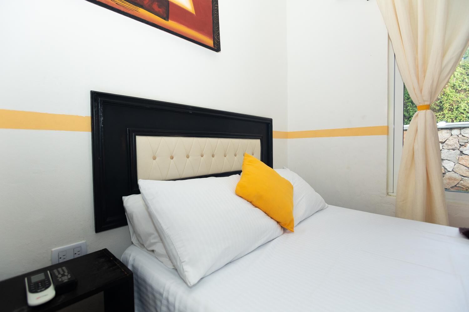 A neatly made bed with white and yellow pillows, a black headboard, and a small bedside table with a phone and a box.