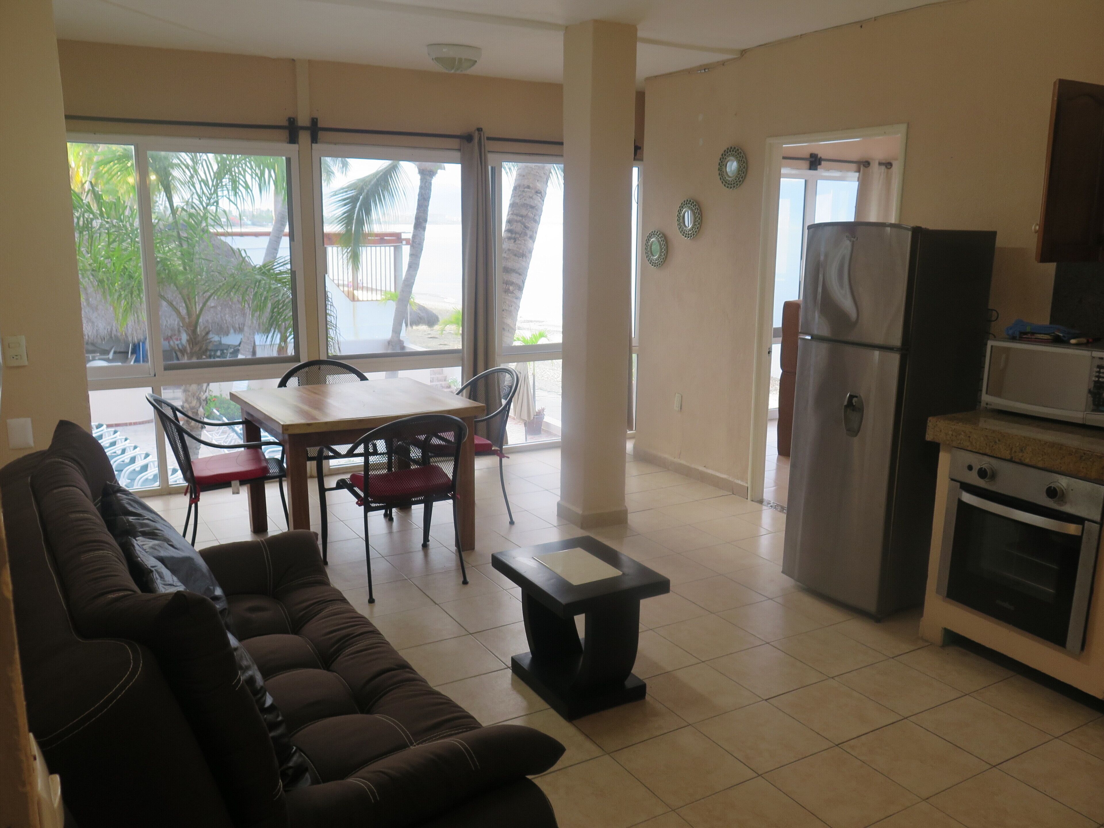 A living room with a sofa, a dining table with chairs, a kitchen area with a refrigerator and microwave, and large windows with a view of palm trees.